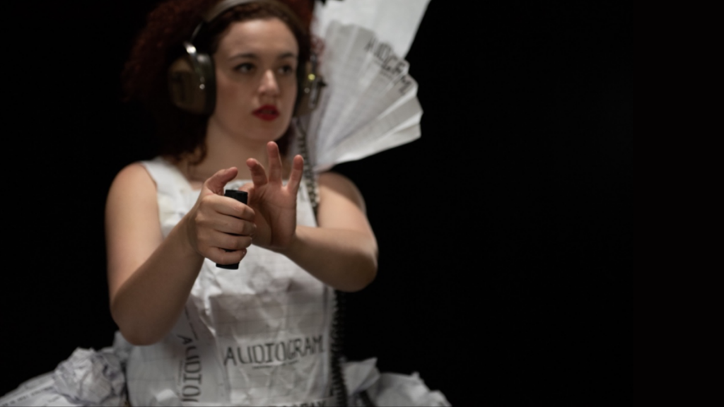 A woman wearing oversized head phones and an elaborate dress made from the white pages of an audiogram, is poises to click a remote trigger that she holds outstretched in front of her