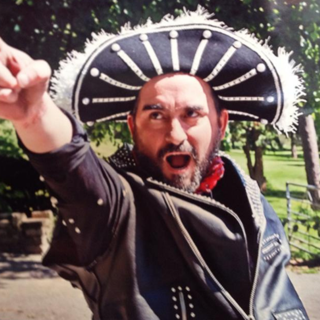 A white man with a beard points towards behind the photographer. He is dressed in a studded leather waistcoat and an OTT feathered bandito style hat