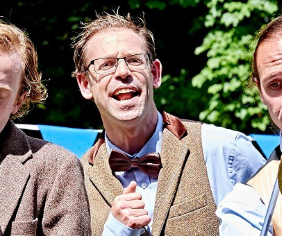 A white man in costume for a show. His mouth is open, possibly in song. He wears a tweet waistcoat, blue shirt and silk bowtie. Two other men flank him, they are mainly cropped out of shot.