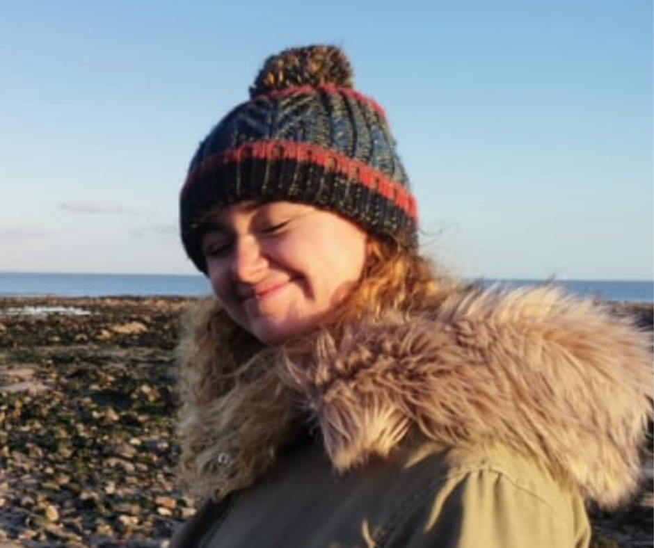 A young white woman wearing a winter coat and bobble hat squints into the camera on a sunny day on the beach