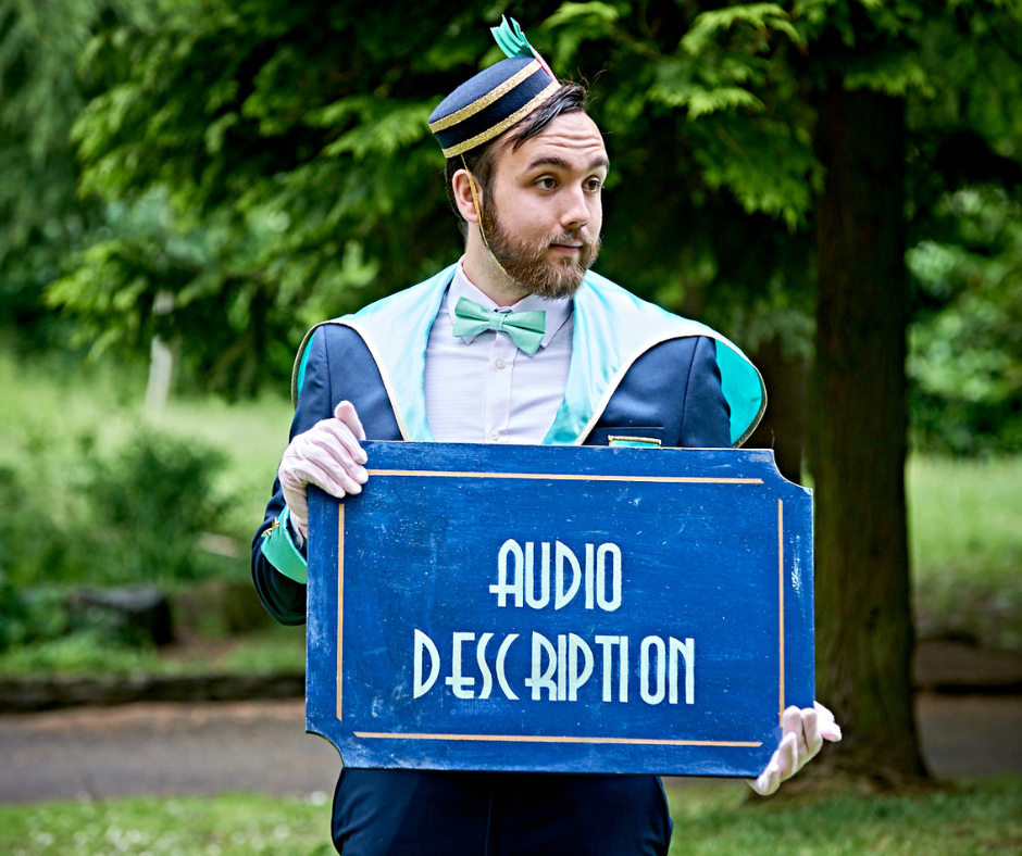 A young whie man with mid brown hair and beard, costued as a 1920s bellhop, he holds a blue board that reads Audio Description