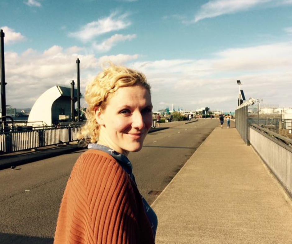 A white woman with blond curly hair looks over her shoulder at the camera, she is outside walking across a bridge on a sunny day