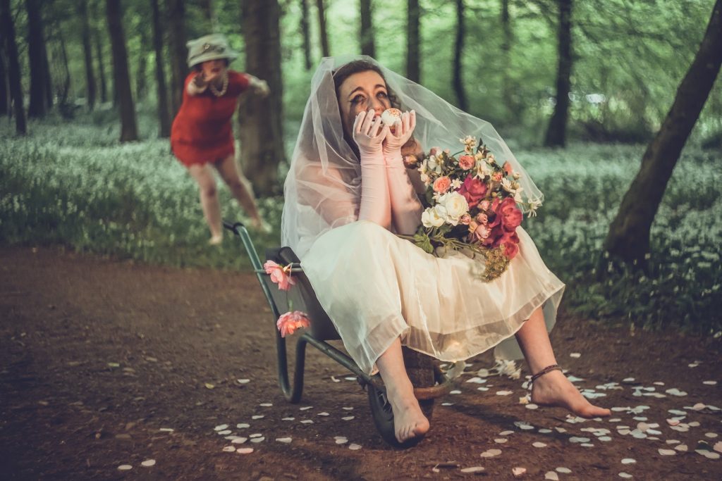 A jilted bride with her bouquet cries in a wheelbarrow in a forest, the Mother of the bride runs after her