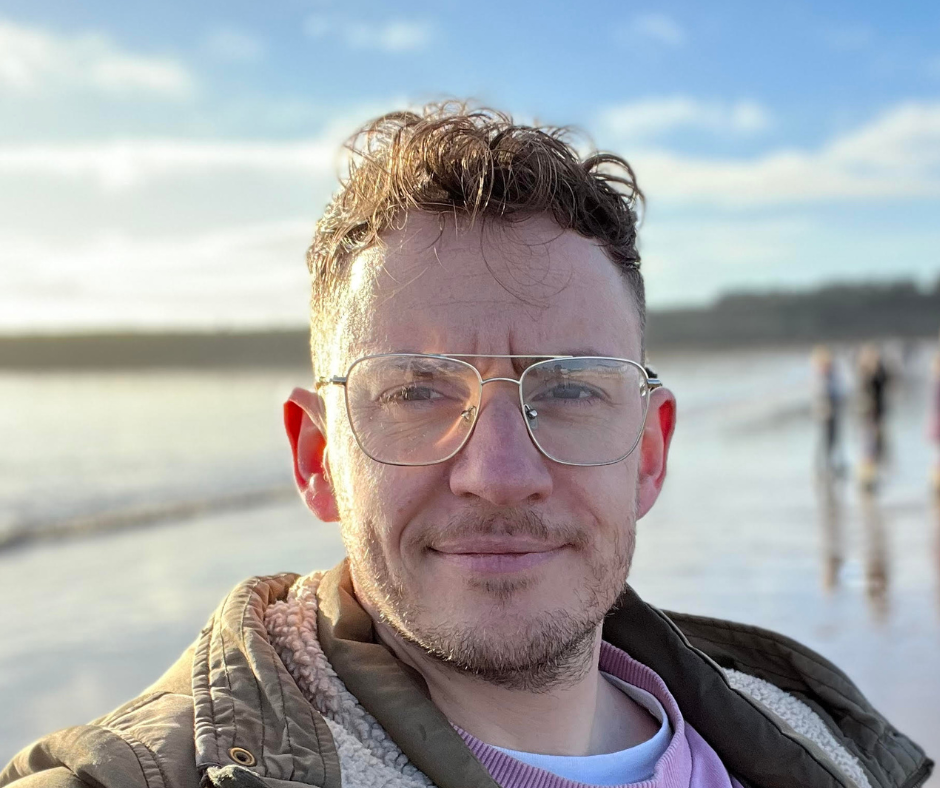 A selfie of a man on a beach. He is white with glasses and stubble, he wears a heavy coat, it is probably a winter's day