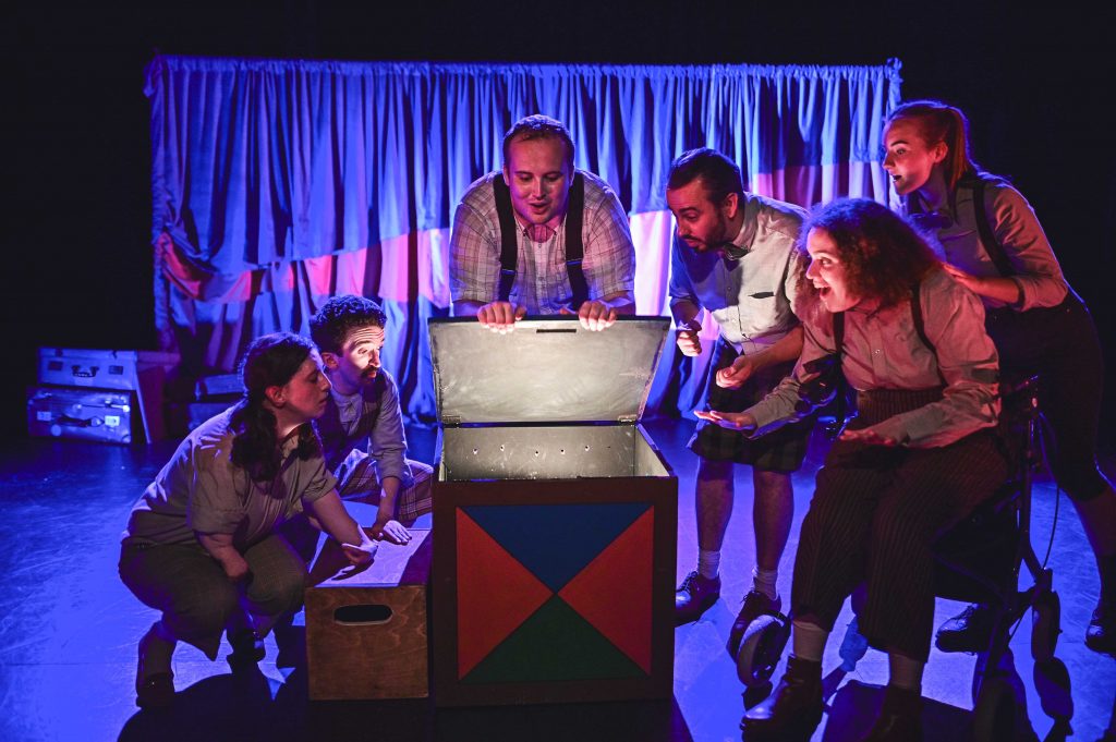 The cast of First Three Drops, costumed in brown and beige hues gather excitedly around a large box, from which escapes a magical light