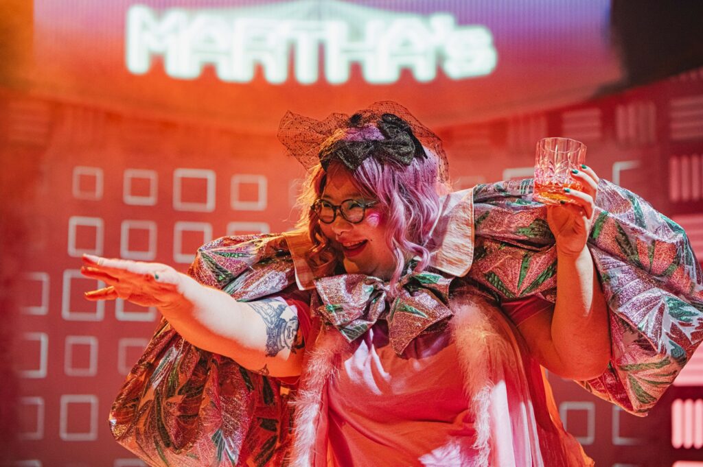 Bea in pink with black rimmed glasses and oversized sleeves smiles broadly and points out of shot, bathed in orange light