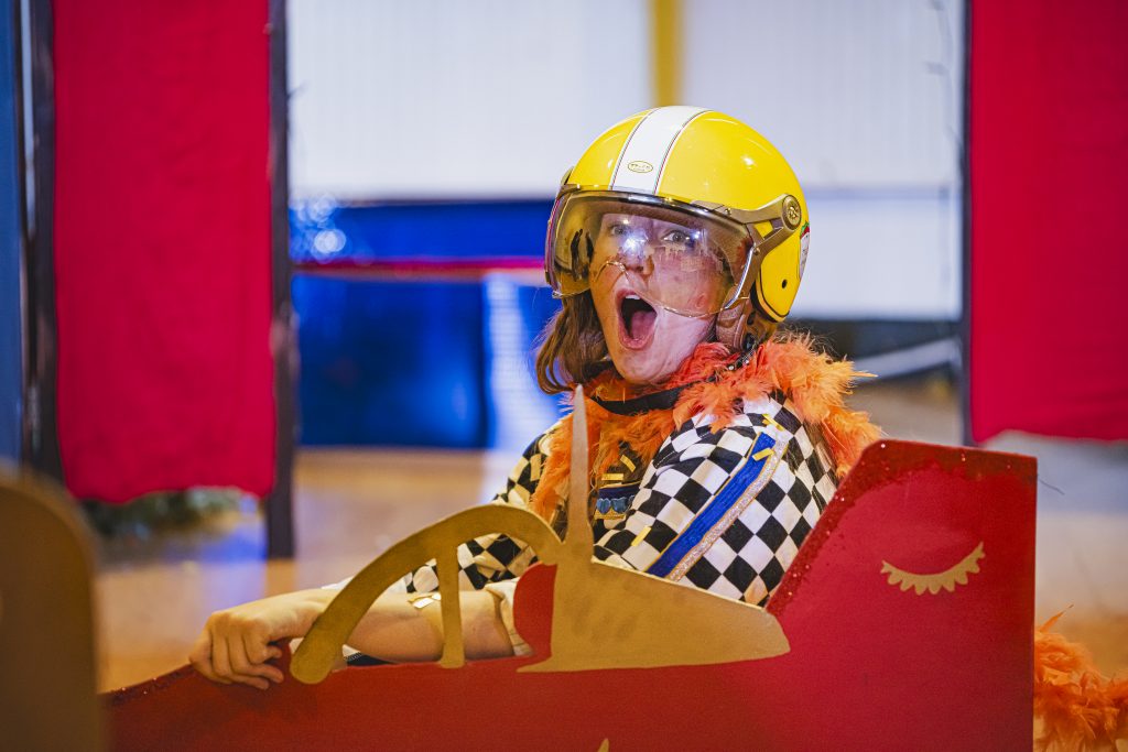 A performer with a wide-eyed, excited expression sits inside a red, flat-board car prop on a stage. They are wearing a bright yellow racing helmet with a clear visor, a black-and-white checkered shirt, and a vibrant orange feather boa. Red curtains and a blue backdrop frame the theatrical scene.