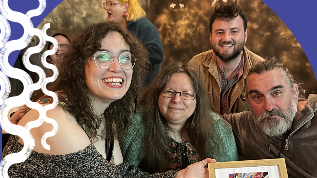 A group of four people posing and smiling together, holding a framed certificate or award. The group includes a person with long, dark curly hair and glasses in the foreground and a man with a grey beard on the right. Two other people are visible in the soft-focus background. The left side of the image features a decorative purple border with white wavy lines.